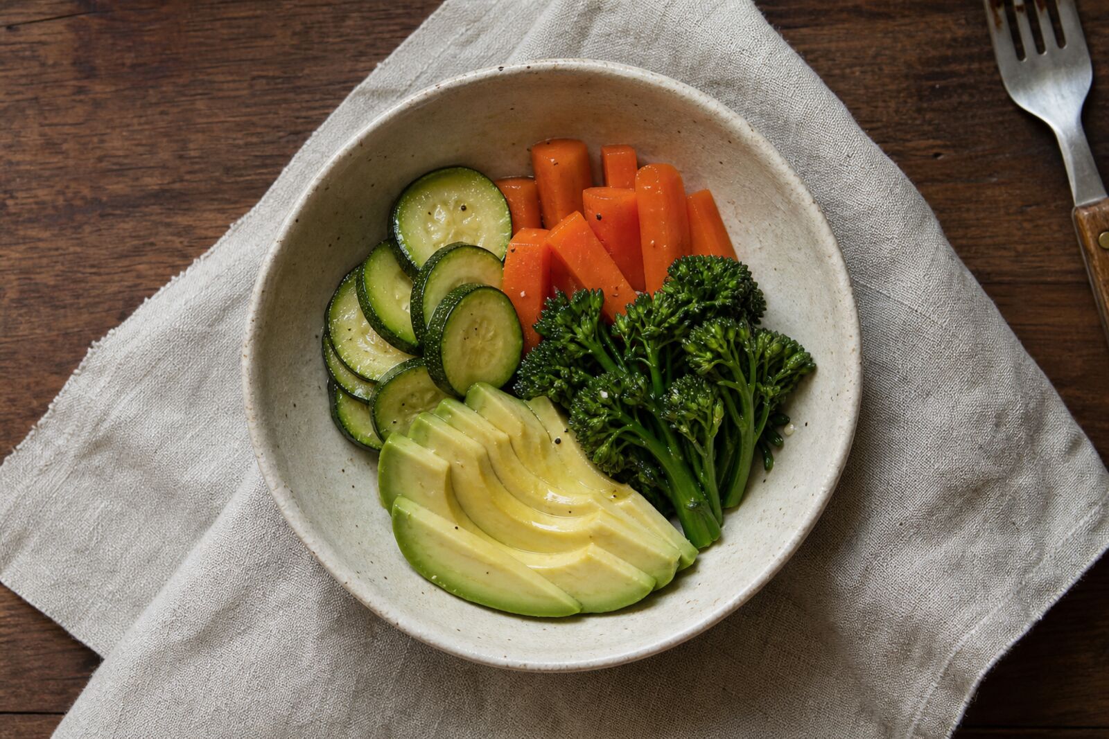 Steamed vegetables and sliced avocado in a shallow bowl on a linen napkin, overhead